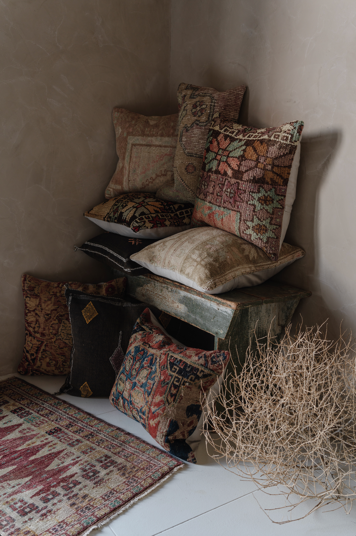 A styled collection of vintage, distressed, and patchwork throw pillows in earthy tones, arranged on a rustic wooden crate in a corner of a room with a textured plaster wall and a patterned rug.