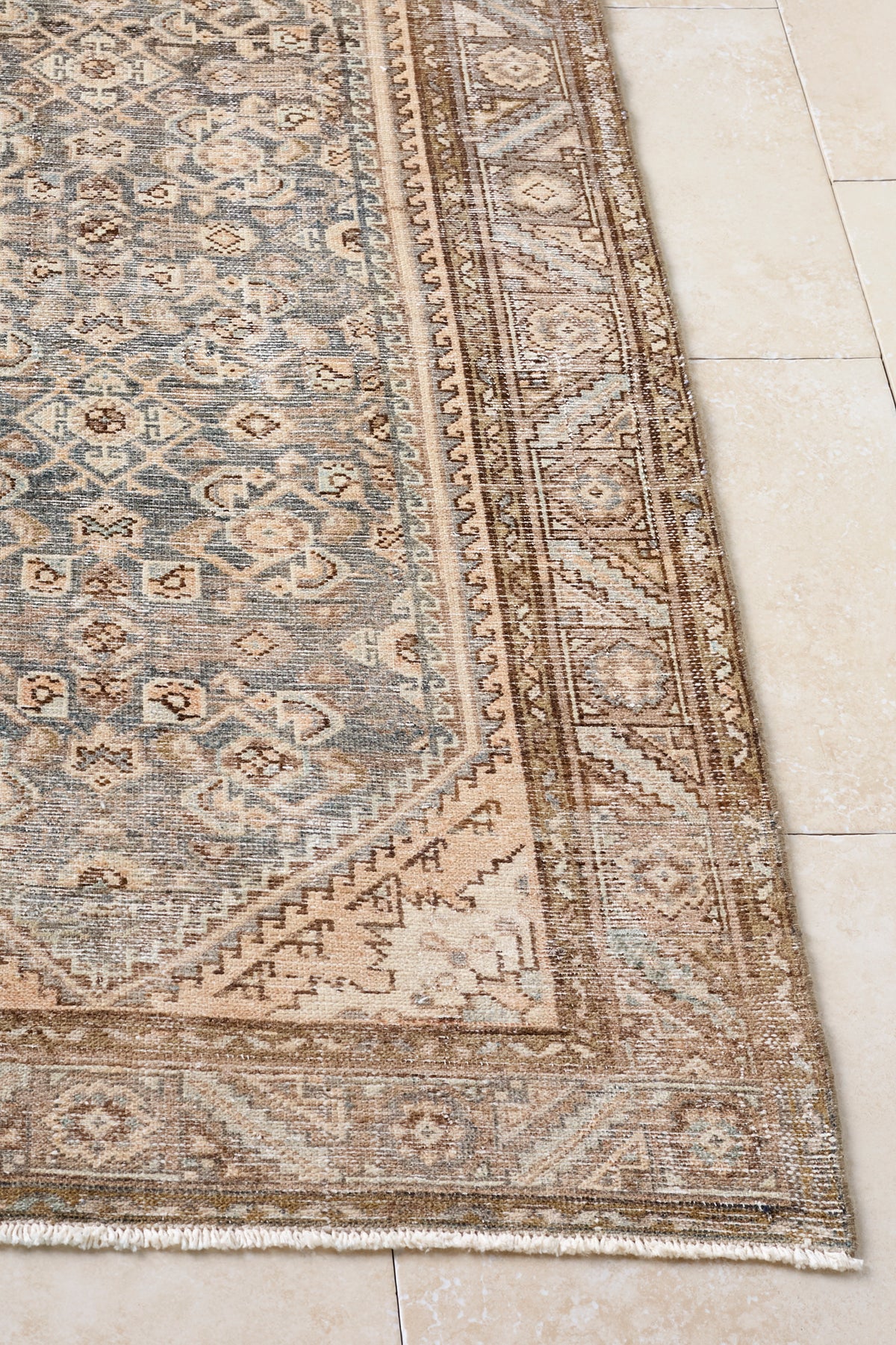 A close-up of a traditional patterned area rug corner, showing a beige and gray-blue design with a detailed border, laid out on a light beige tiled floor.